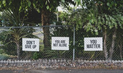 A fence in front of lush green holding up signs that say "Don't Give Up," "You Are Not Alone," and "You Matter."
